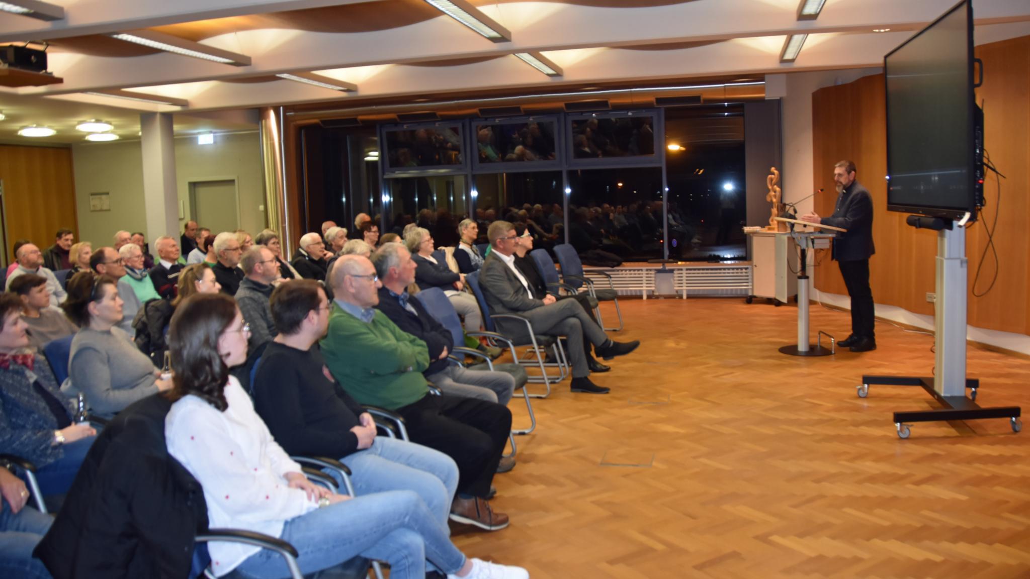 Etwa 80 Menschen sitzen auf Stühlen und hören den Worten des Dekans zu. Neben dem Dekan steht ein großes Fernsehgerät